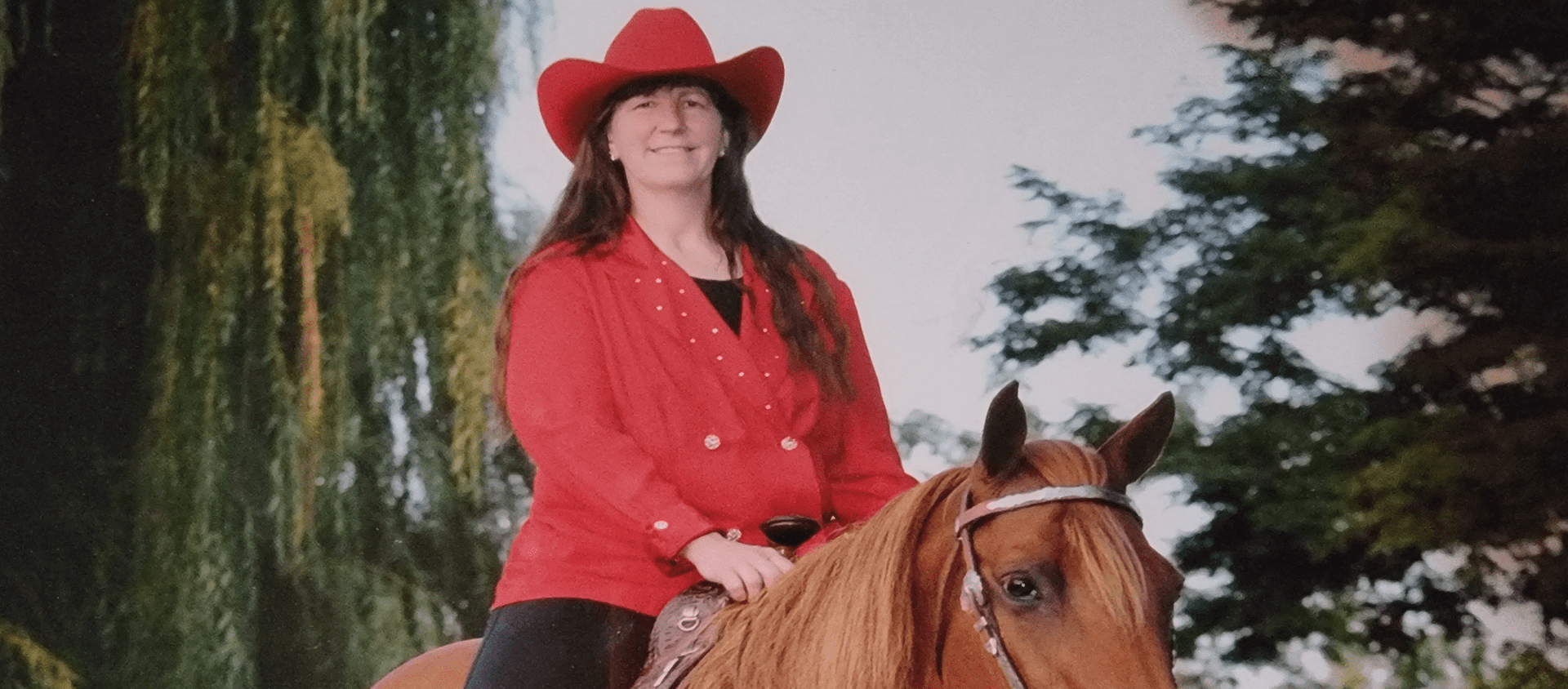 Woman in red coat and hat riding a brown horse outdoors.